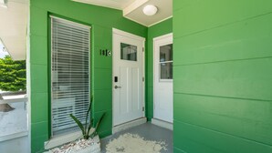 A green house exterior with a white door, a window with blinds, and a small planter with rocks. - Dune Your Thing C (Fort Myers Beach)