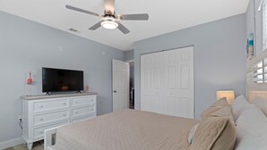 Ein Schlafzimmer mit Bett, Fernseher, Kommode, Deckenventilator und einer Tür.