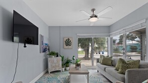 Una sala de estar con sofá, mesa de centro y plantas, un ventilador de techo y un televisor montado en la pared.