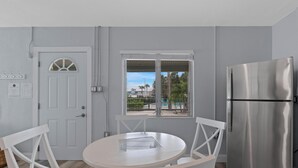 Una cocina moderna con un refrigerador de acero inoxidable, una mesa de comedor blanca con sillas, una puerta blanca con ventana y vistas a una piscina y palmeras afuera.