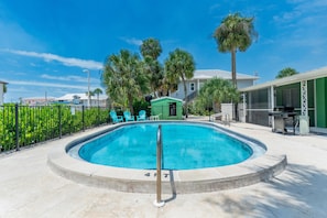 A swimming pool with a metal safety rail, surrounded by a fence, palm trees, and a building with a green exterior.