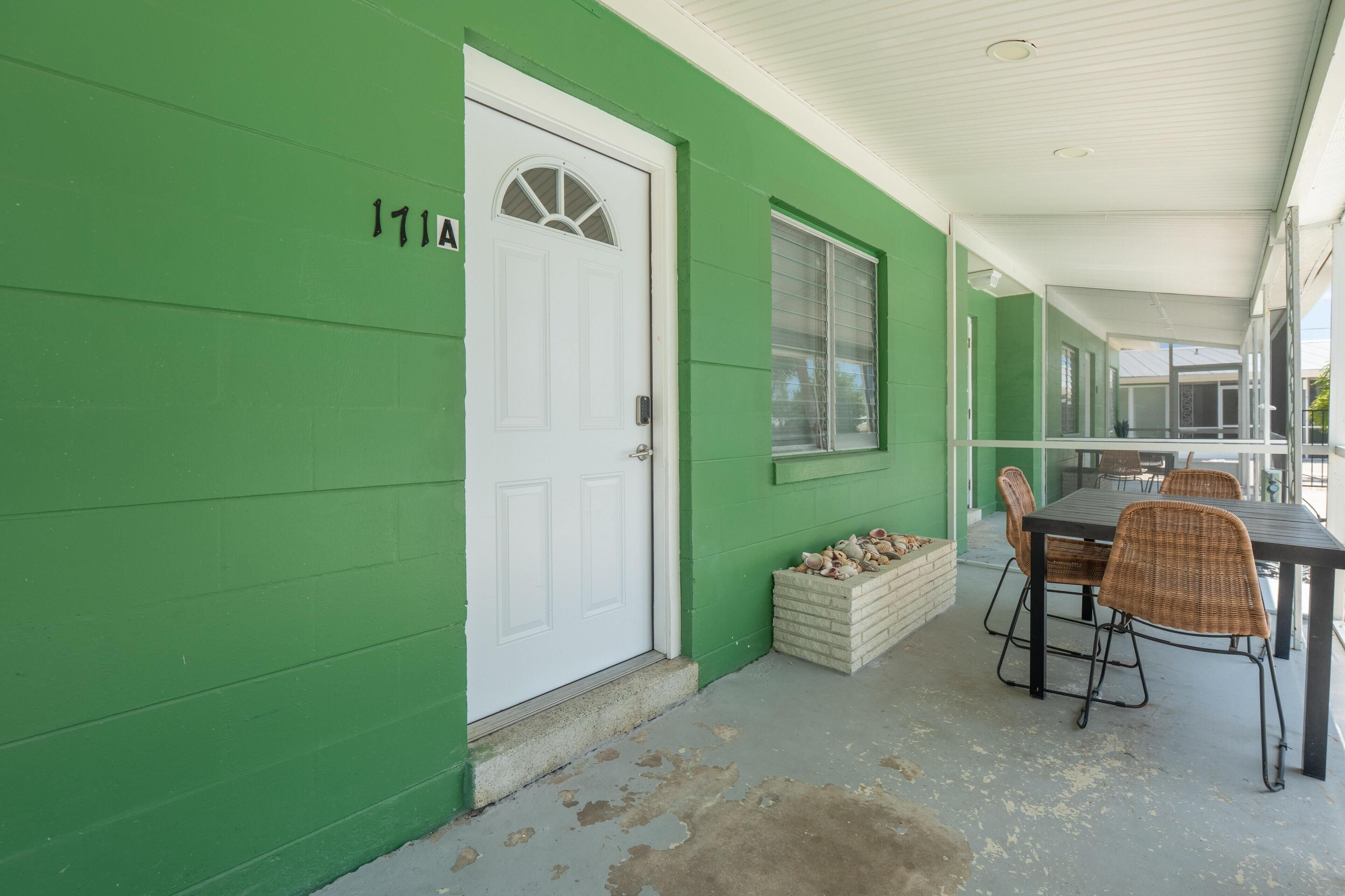 Un bâtiment vert avec une porte blanche, une fenêtre et une table entourée de chaises à l’extérieur.