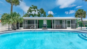 A swimming pool with a covered area, palm trees, and a clear sky. - Dune Your Thing A (Fort Myers Beach)