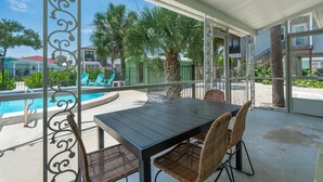 A covered outdoor area with a pool, a table with chairs, and a view of the surrounding buildings and greenery. - Dune Your Thing A (Fort Myers Beach)