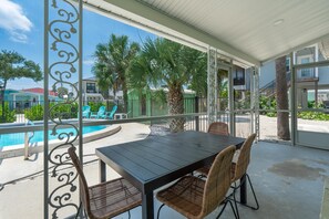 A covered outdoor area with a pool, a table with chairs, and a view of the surrounding buildings and greenery. - Dune Your Thing A (Fort Myers Beach)
