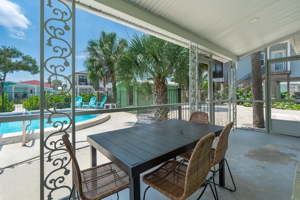A covered outdoor area with a pool, a table with chairs, and a view of the surrounding buildings and greenery. - Dune Your Thing A (Fort Myers Beach)