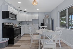 A modern kitchen with white cabinetry, a black stove, a dining table with white chairs, and a view of the outdoors through a large window. - Dune Your Thing A (Fort Myers Beach)