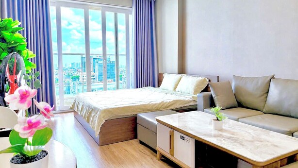 A modern hotel room with a bed, sofa, and dining table, large windows with curtains, and a city view. - SKYHOME Apartment (Ho Chi Minh City)