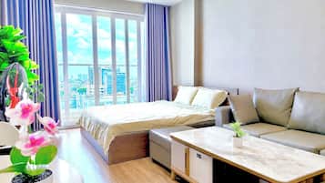 Una habitación de hotel moderna con cama, sofá y mesa de comedor, ventanas amplias con cortinas y vistas a la ciudad.