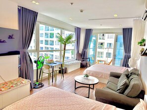 Ein modernes Hotelzimmer mit zwei Betten, einem Esstisch, einem Stuhl, einem kleinen runden Tisch und einem großen Fenster mit Vorhängen.