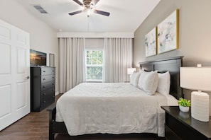 Ein Schlafzimmer mit Bett, Kommode, Nachttisch, Deckenventilator, Fenster mit Vorhängen und Kunstwerken an der Wand.
