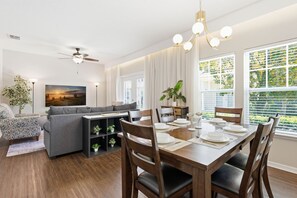 Ein modernes Wohnzimmer mit Essbereich, Deckenventilator und großen Fenstern mit Jalousien.