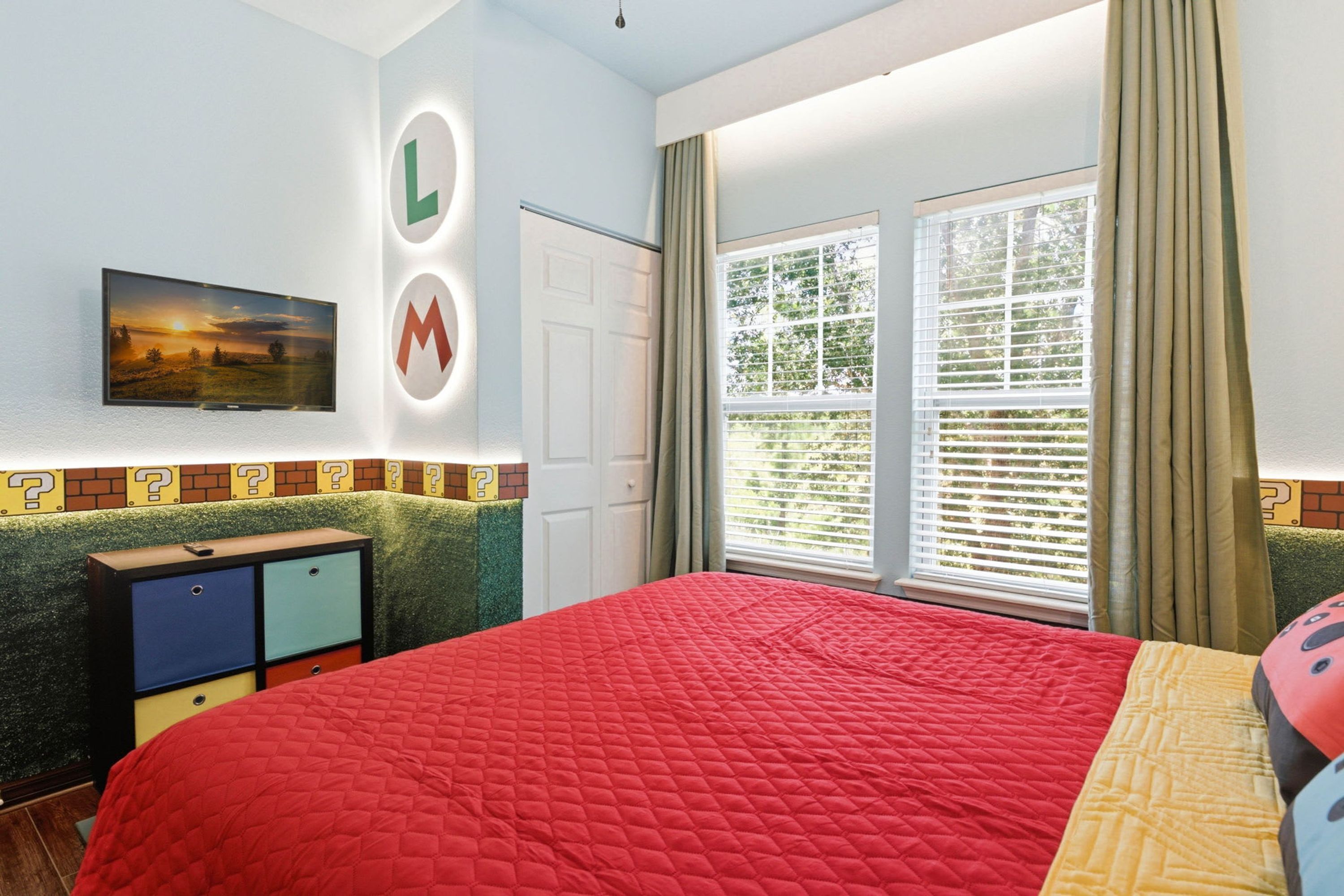 A bedroom with a red quilted bedspread, a television on the wall, a dresser, and a window with curtains.