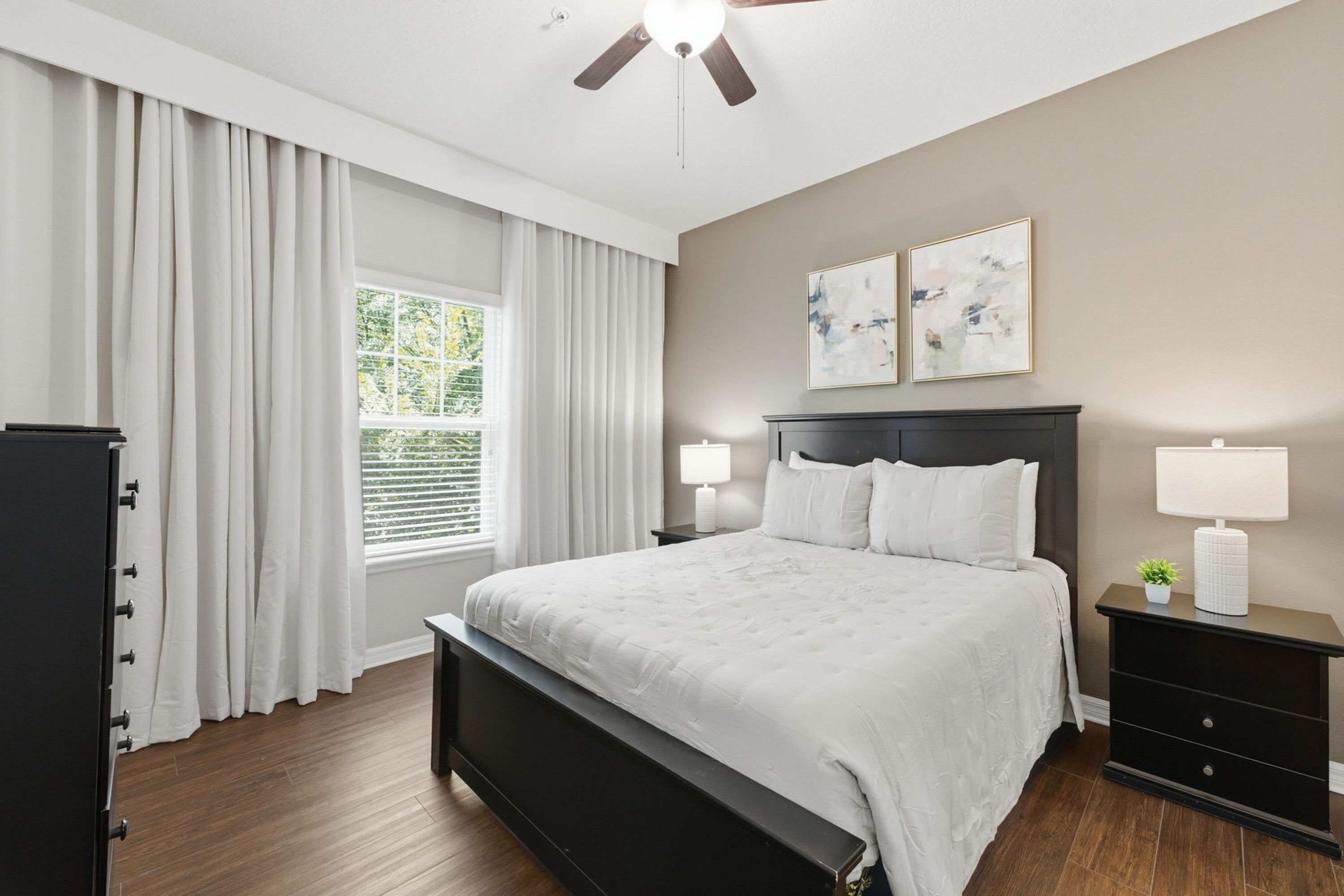 A bedroom with a bed, a nightstand, a dresser, a ceiling fan, and a window with curtains.