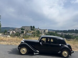 Ein altes, schwarzes Auto steht an einer Straße, im Hintergrund ein historischer Gebäudekomplex.