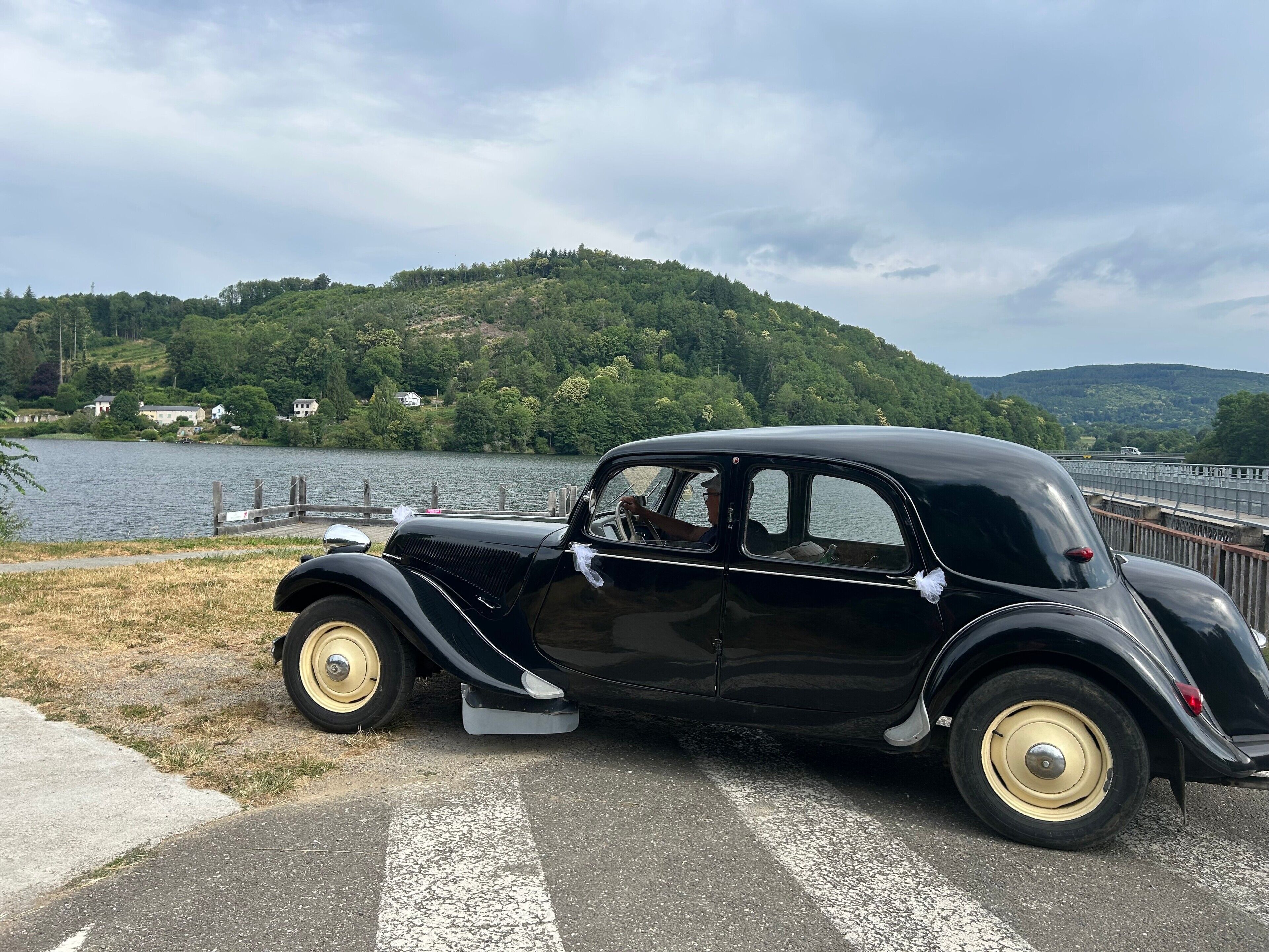 Ein klassisches schwarzes Auto parkt am Seeufer, im Hintergrund ein bewaldeter Hügel.