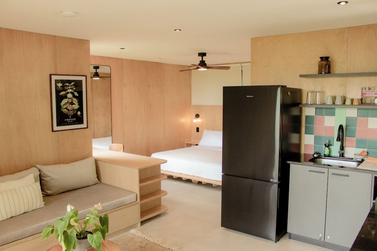 Una cocina compacta con refrigerador, una zona de comedor con mesa y sillas, y una cama al fondo.