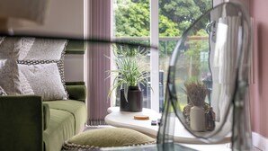 Een kamer met een groene bank, een kleine tafel met een potplant, een spiegel en een raam met uitzicht op bomen.