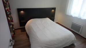 A hotel room with a bed, a headboard, a window with curtains, a radiator, and a door with a handle.