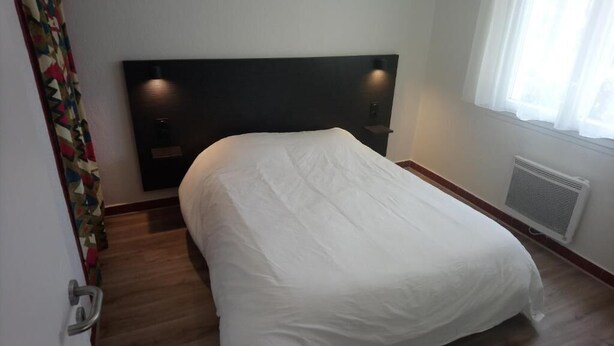 A hotel room with a bed, a headboard, a window with curtains, a radiator, and a door with a handle.