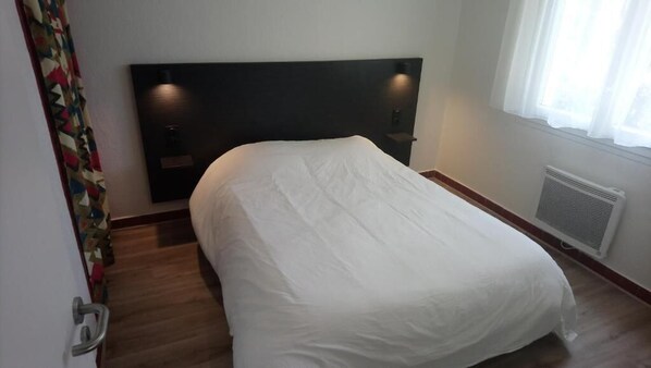 A hotel room with a bed, a headboard, a window with curtains, a radiator, and a door with a handle. - Le Vendahaut Résidence de tourisme (Lapleau)