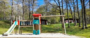 A playground with a slide, climbing structures, and a swing set in a wooded area.