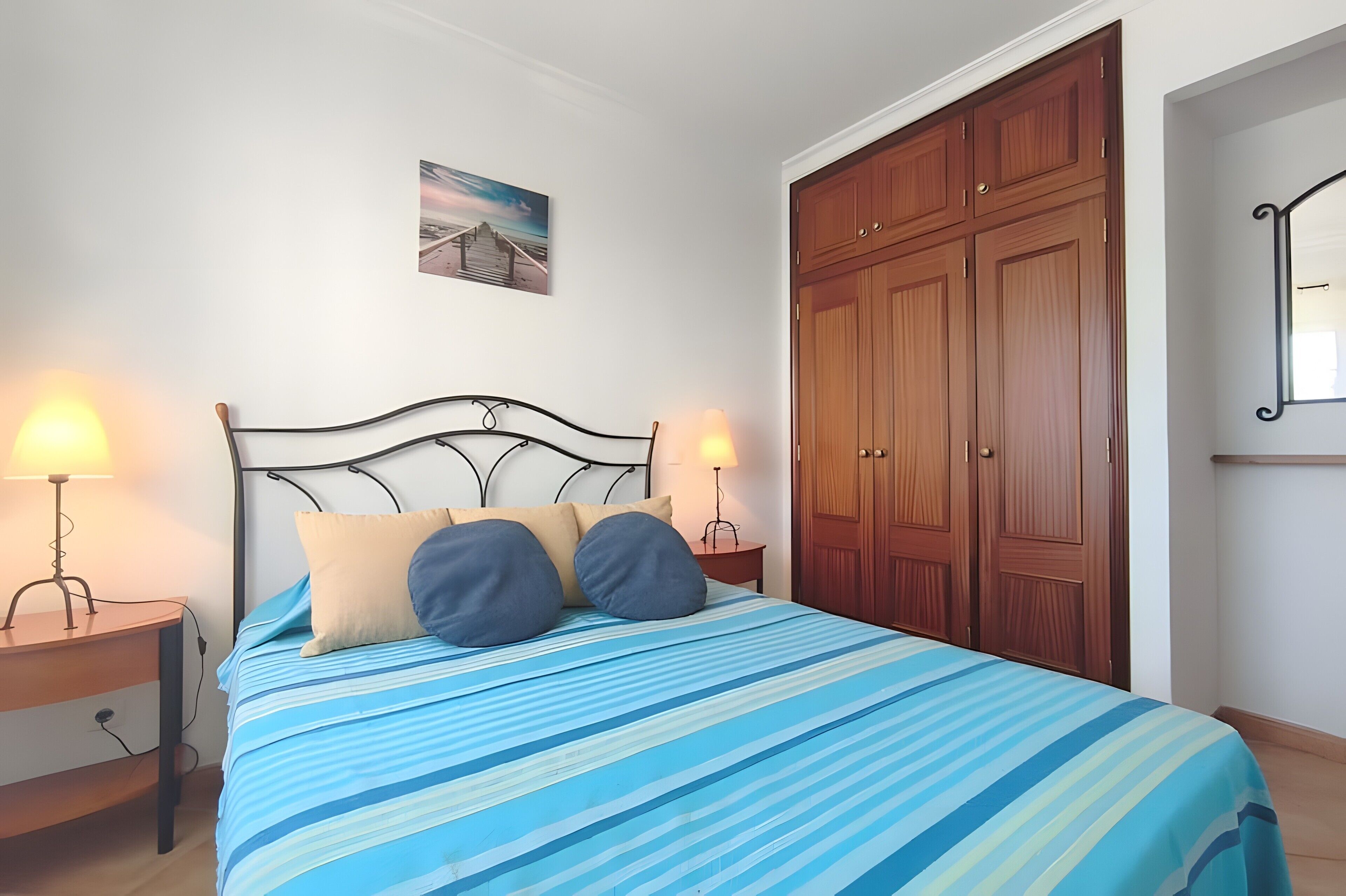 A bedroom with a bed, two bedside lamps, a wooden wardrobe, and a framed picture on the wall.