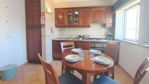 Een kleine keuken met houten kastjes, een eettafel voor vier personen en een raam dat voor natuurlijk licht zorgt.