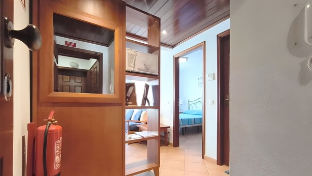 Een gang met een brandblusser, een houten deur en een blik op een kamer met een bed en bureau.
