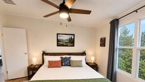 Ein Schlafzimmer mit einem Bett, einem Deckenventilator, einem Fenster mit Vorhängen, einem Bild an der Wand und einem Nachttisch mit Lampe.