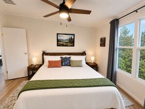 Ein Schlafzimmer mit einem Bett, einem Deckenventilator, einem Fenster mit Vorhängen, einem Bild an der Wand und einem Nachttisch mit Lampe.