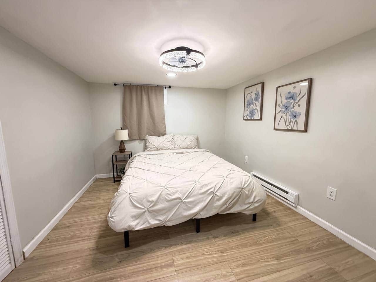 Une chambre à coucher comprenant un lit, une table de chevet, une lampe, un ventilateur de plafond et deux tableaux accrochés au mur.