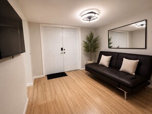 Una habitación con un sofá de cuero negro, una puerta blanca, una pequeña planta en maceta, un ventilador de techo y un televisor montado.