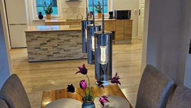 Ein moderner Essbereich mit einem Holztisch, zwei Stühlen, einer Vase mit Blumen und einer Küche im Hintergrund.