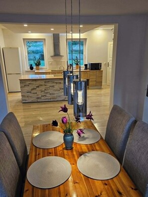 A modern dining area with a wooden table, two chairs, a vase with flowers, and a kitchen in the background.