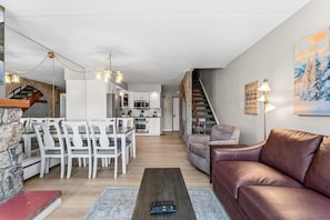 Ein modernes Wohnzimmer mit einem braunen Ledersofa, weißen Esstischen und einem Holztisch. Im Hintergrund ist eine Küche zu sehen.