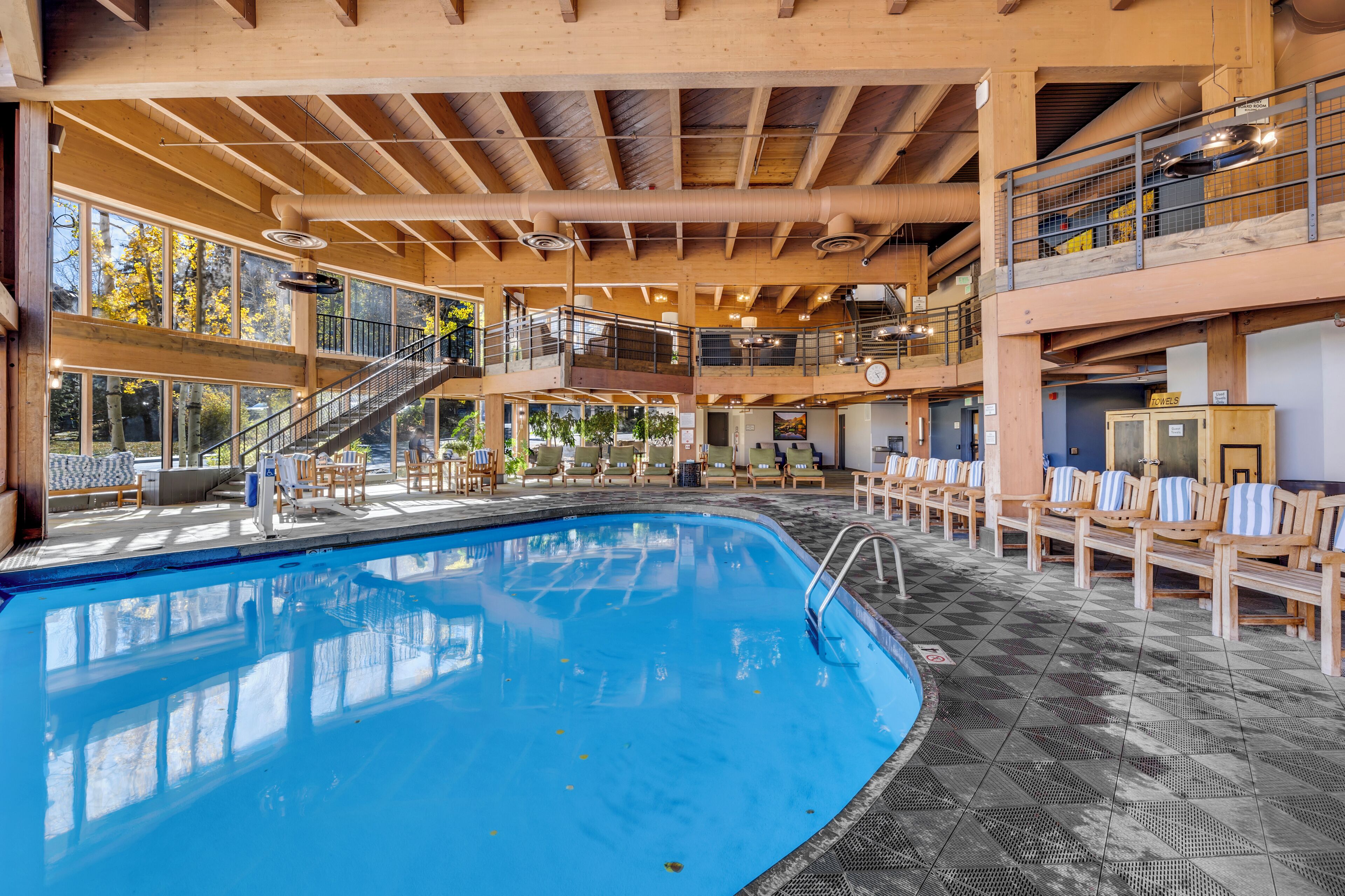 A large indoor swimming pool with a wooden deck area, multiple levels with railings, and a view of trees outside.