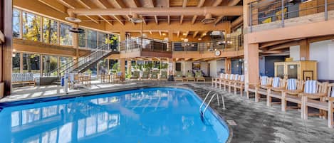 A large indoor swimming pool with a wooden deck area, multiple levels with railings, and a view of trees outside.