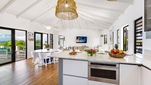 A modern kitchen with a central island, a dining area with white furniture, and a large window with a view. - Peace & Happiness Anguilla (Cul du Sac)