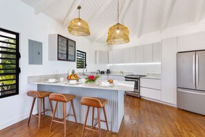 A modern kitchen with a central island, bar stools, pendant lights, and stainless steel appliances. - Peace & Happiness Anguilla (Cul du Sac)