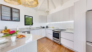 A modern kitchen with white cabinetry, a central island, a built-in oven, and a large window with greenery outside. - Peace & Happiness Anguilla (Cul du Sac)