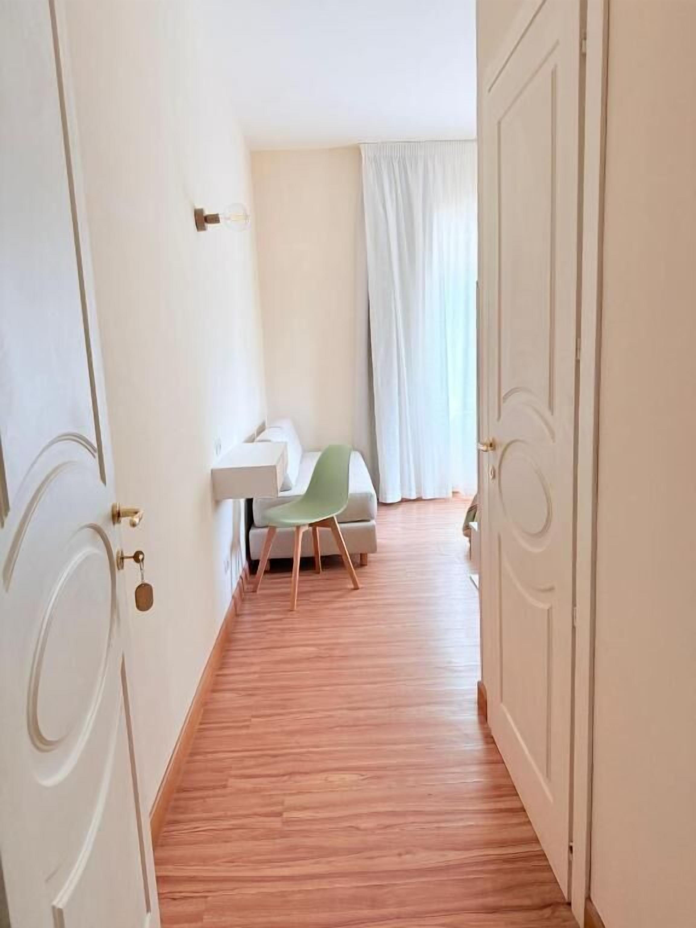 Un pasillo con suelo de madera, una puerta blanca a la izquierda, una pared blanca con un interruptor de luz y una silla verde al fondo.