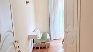 Käytävän lattia on puuta, vasemmalla puolella on valkoinen ovi, seinä on valkoinen ja siinä on valokytkin, ja kauempana näkyy vihreä tuoli.