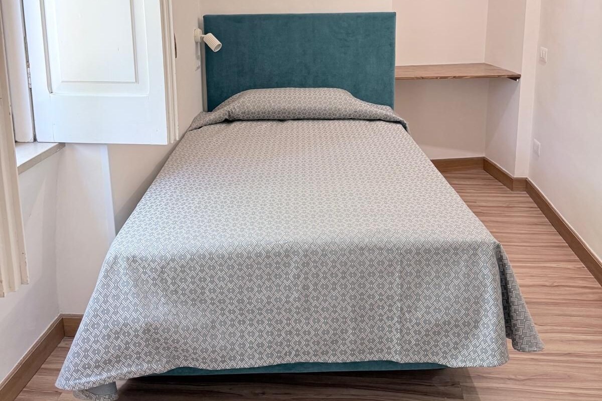 Una habitación pequeña con cama individual, cabecera color turquesa, colcha estampada y piso de madera.