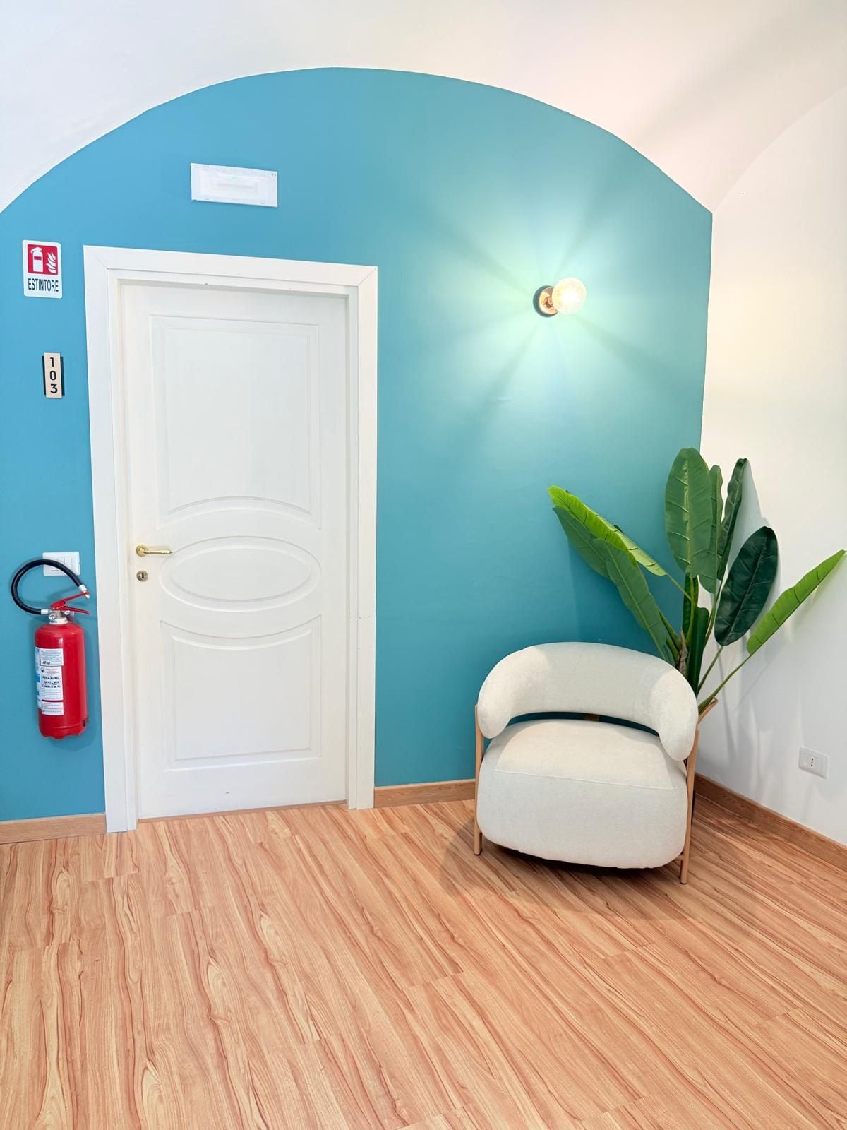 Una stanza con una porta bianca, un estintore, una pianta in vaso e un lampadario a parete.