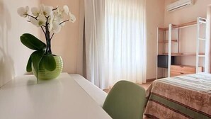 Huoneessa on sänky, työpöytä, tuoli, ikkuna, jossa on verhot, ja seinälle asennettu ilmastointilaite.