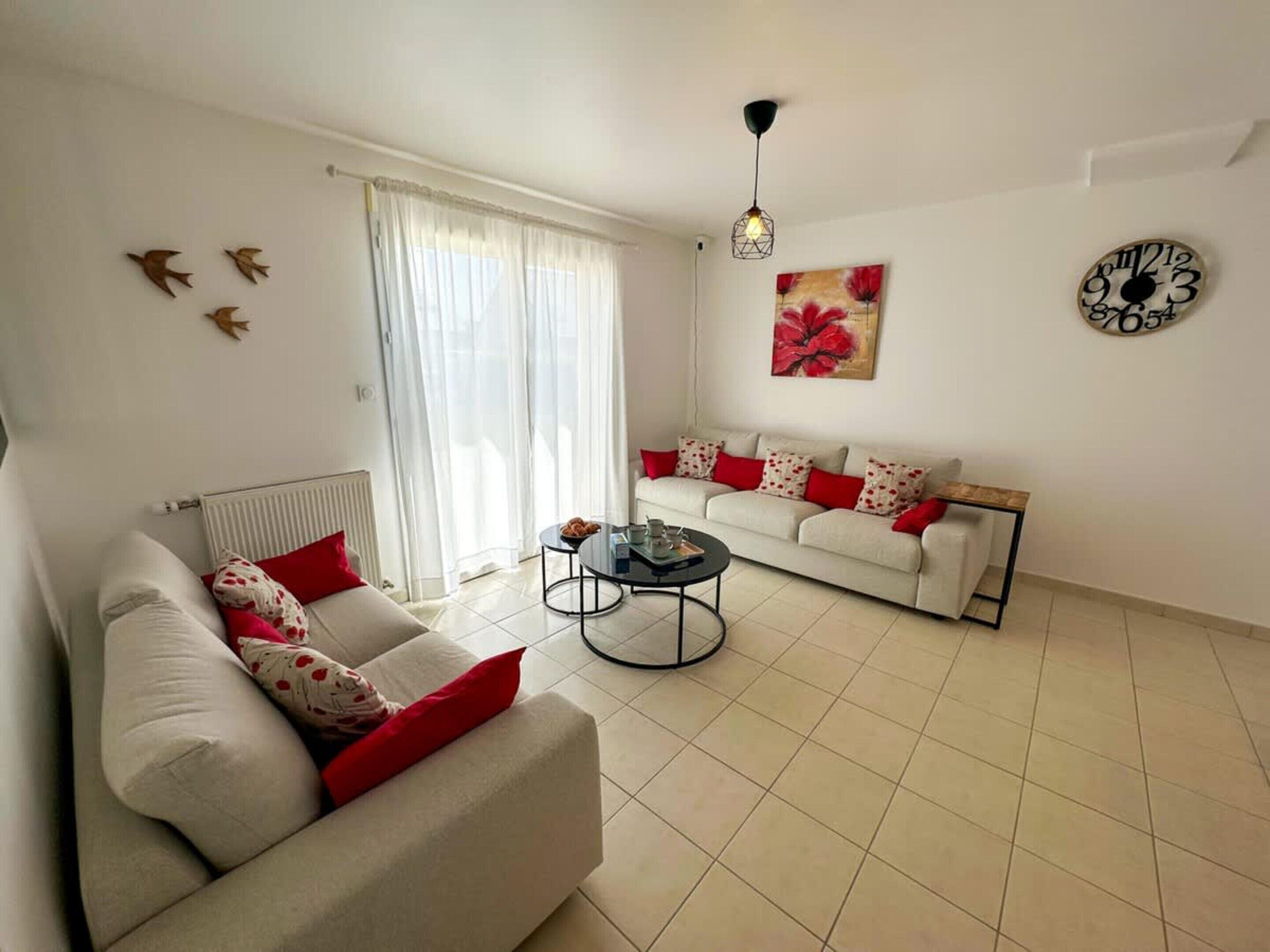 Una sala de estar con un sofá, una mesa de centro, un radiador, una ventana con cortinas, un reloj de pared y un accesorio de iluminación en el techo.