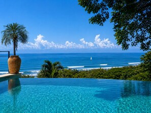 A clear view of an infinity pool with a palm tree and a large vase, overlooking the ocean with a sailboat. - Vista Naranja Luxury Villa - Santa Teresa (Santa Teresa)