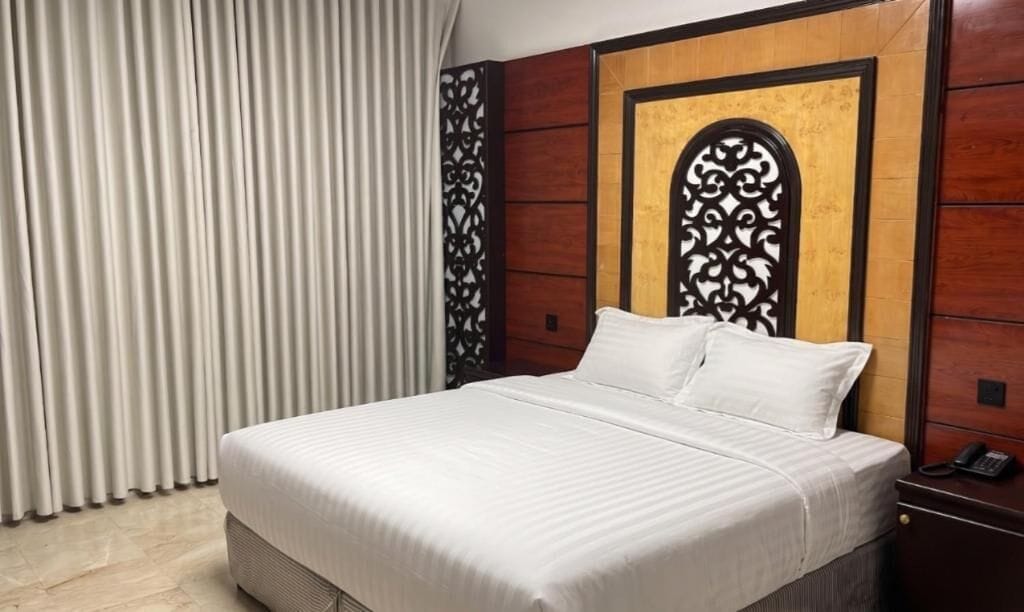 Une chambre d’hôtel avec un grand lit, une tête de lit en bois, une table de chevet avec un téléphone et un rideau.