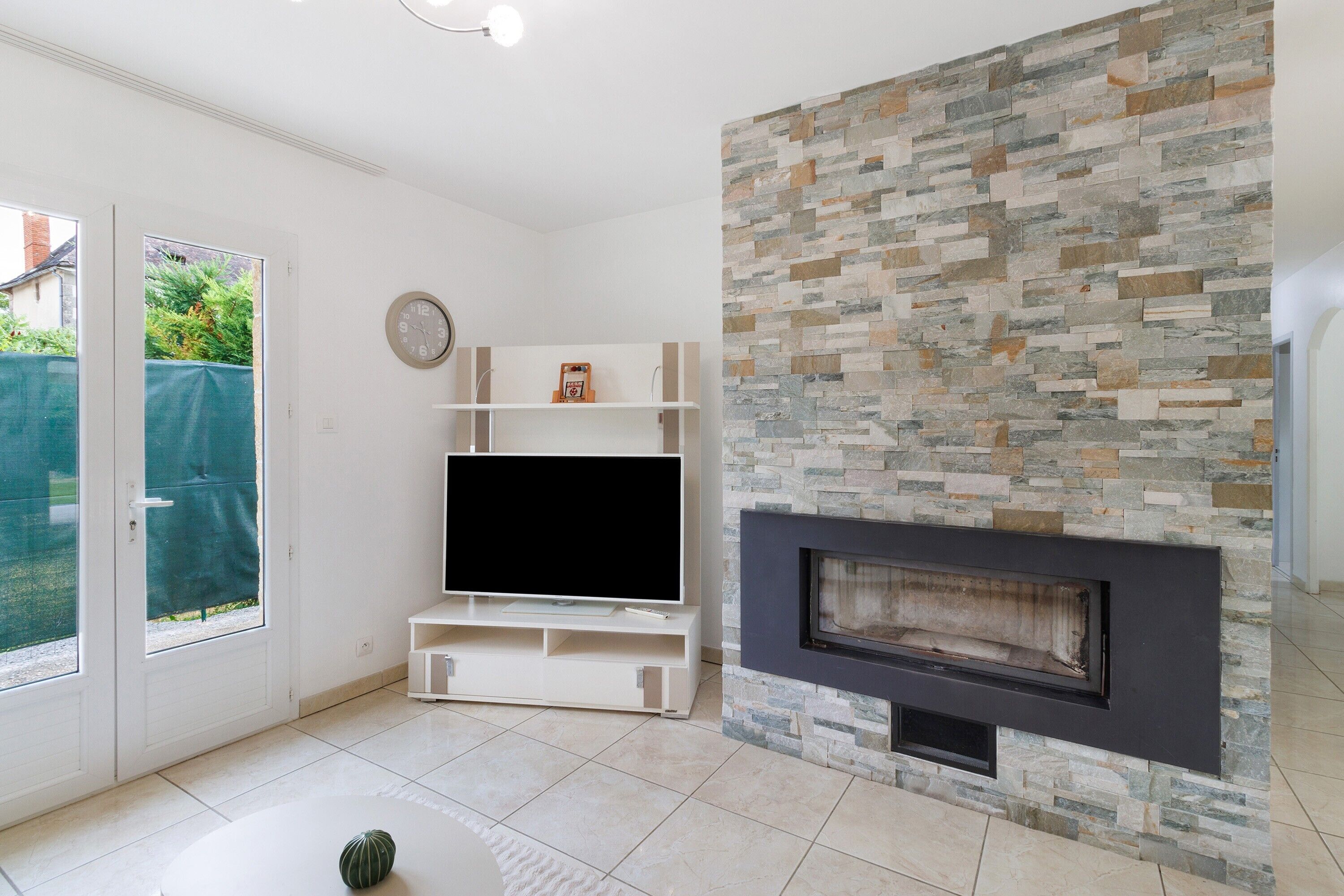 Een woonkamer met een stenen openhaard, een flatscreen televisie op een witte tv-meubel en een schuifpui die uitkomt op een tuin.